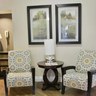 A cozy living room corner featuring two patterned chairs, a round wooden table with a lamp, and framed artwork on the wall.