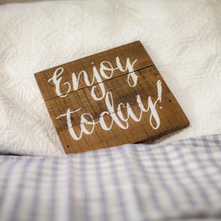 A wooden sign with the message 'Enjoy today!' rests on cozy bedding.