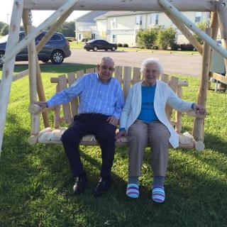 An elderly couple sitting together on a wooden swing in a sunny park.