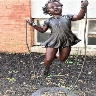 A bronze statue of a girl joyfully jumping rope outdoors.