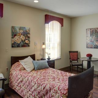 A cozy bedroom with a single bed adorned with a patterned red comforter, two pillows, and flanked by artwork on the walls. The room features a small table with chairs and a window allowing natural light.