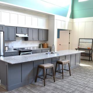 A spacious modern kitchen featuring a large island counter with stools, gray cabinets, and stainless steel appliances. The space is illuminated by natural light pouring through a window near the light blue ceiling.