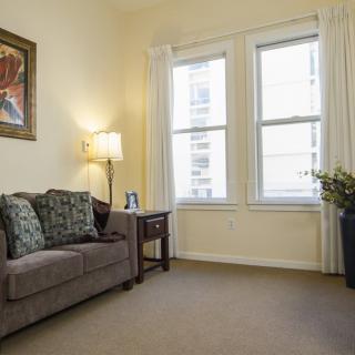 A cozy living room with a comfortable sofa, a side table with a lamp, large windows, and an indoor plant.