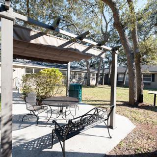 A sunny outdoor patio area with a pergola, table, and chairs surrounded by trees.