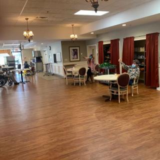 A spacious dining area with hardwood floors, tables, and chairs, illuminated by ceiling lights and natural light from windows.