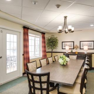 A spacious conference or dining room with a long table and several chairs, illuminated by a chandelier and decorated with artwork and plants.