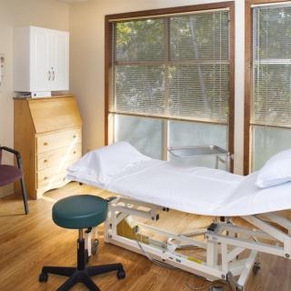 A clean and bright medical examination room with a table, chair, and windows.