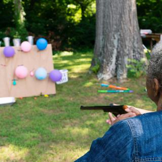 A person aims a pistol at a backyard target setup with colorful balloons.