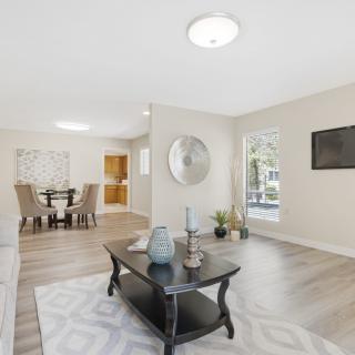 A modern and spacious living room with contemporary furniture and bright lighting, featuring wood flooring and a dining area in the background.