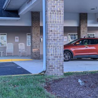 A red car parked in a covered area of a brick building, with a sidewalk and grass in view.