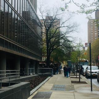 A bustling urban scene featuring pedestrians on a sidewalk next to modern buildings and greenery.