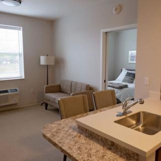 A view of a modern apartment with an open living area, kitchen counter, and cozy bedroom.
