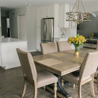 A modern dining area with a wooden table and cushioned chairs, featuring a stylish chandelier and a vase of yellow flowers.