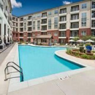 A modern apartment complex featuring a large outdoor swimming pool surrounded by patio furniture on a sunny day.