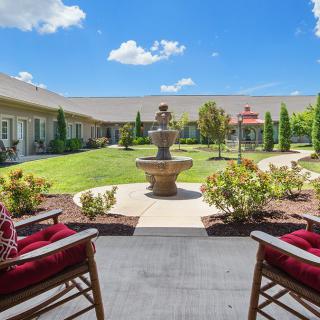 A tranquil courtyard featuring a central fountain, surrounded by well-maintained gardens and comfortable outdoor seating under a clear blue sky.
