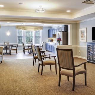 A spacious and well-lit seating area with several chairs and tables, featuring a carpeted floor and large windows.