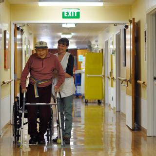 An elderly person using a walker is assisted by a caregiver in a nursing home hallway.