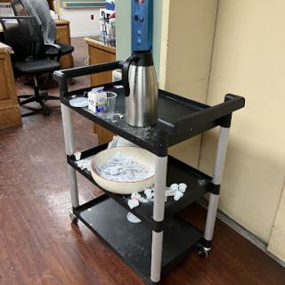 A utility cart in an office setting, featuring a coffee thermos and dishware.