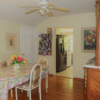 A cozy dining room with a floral-tablecloth-covered table and chairs, adorned with fresh flowers, artwork on the walls, and a view into the kitchen.