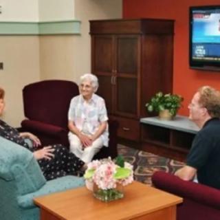 Three people are sitting in a living room, engaged in conversation with a television on the wall.