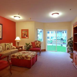 A cozy living room with warm red accents, featuring a sofa, armchair, lamp, and bookshelf, illuminated by natural light from large windows.