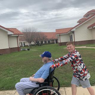 A child joyfully pushes an elderly person in a wheelchair outdoors.