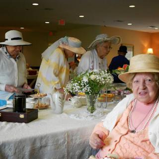 A joyful gathering of elderly women enjoying a tea party, dressed in hats and surrounded by flowers.