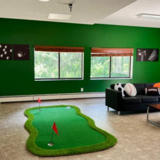 A bright recreation room with a mini golf setup, featuring green walls and large windows.