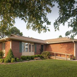 A suburban brick house with a front ramp and lush green lawn surrounded by trees.