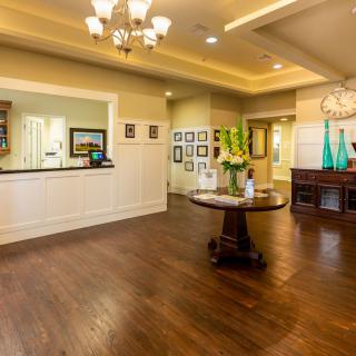 A warmly lit lobby with wooden flooring, featuring a reception desk and decorative elements like a chandelier and flower arrangements.