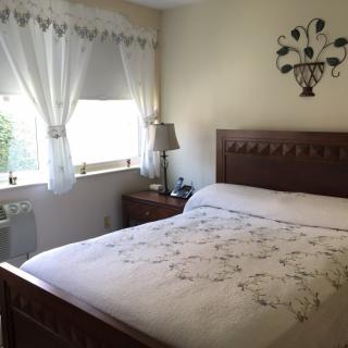 A cozy bedroom with a neatly made bed, a window with white curtains, and a nightstand with a lamp.