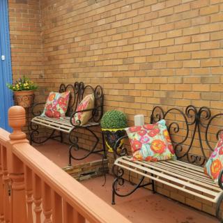 A cozy patio with wrought iron benches adorned with colorful pillows, set against a brick wall, featuring potted plants for decoration.
