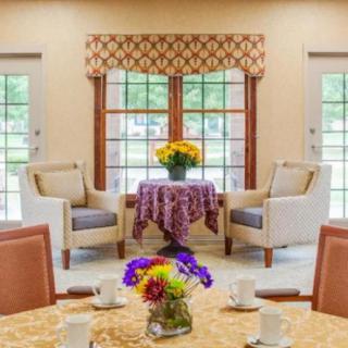 A cozy living room setup with two armchairs and a small table adorned with flowers, framed by large windows and patterned curtains.