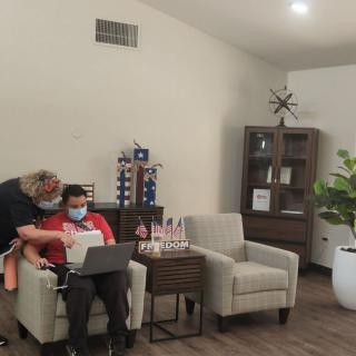 Two people wearing masks are sitting indoors, using a laptop in a cozy room with chairs and a plant.