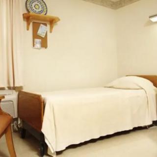 A minimalist hospital room featuring a single bed with white bedding, a chair, and a small shelf on the wall.
