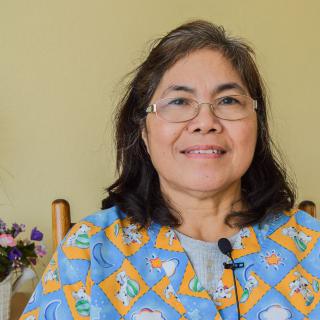 A smiling woman wearing glasses sits indoors with a floral arrangement in the background.