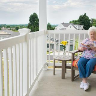 An elderly woman relaxes on a porch, enjoying a book with a view of a residential neighborhood.