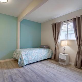 A minimalist bedroom featuring a single bed with a blue accent wall, nightstand with a lamp, and large window with curtains.