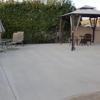 A backyard patio with a gazebo and outdoor furniture set on a concrete surface, surrounded by greenery.