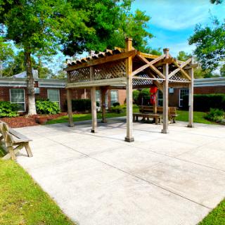 A sunny outdoor courtyard featuring a wooden pavilion, benches, and lush greenery.