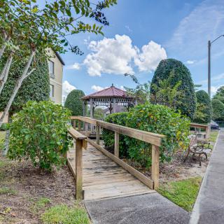 A serene garden scene featuring a wooden bridge leading to a gazebo, surrounded by trees and bushes under a partly cloudy sky.