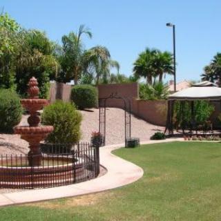 A landscaped garden featuring a fountain, gazebo, and paved pathway surrounded by trees and grass.