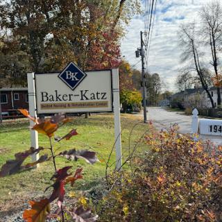 A welcoming sign for Baker-Katz along a scenic road surrounded by autumn foliage.