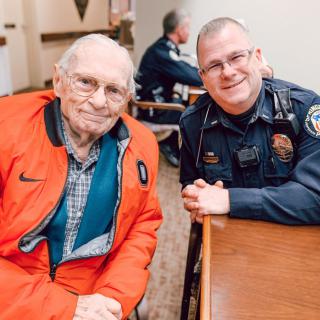 An elderly man and a police officer smiling together indoors, showcasing community interaction.