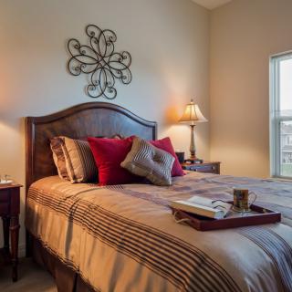 A cozy bedroom with a neatly made bed, adorned with decorative pillows and a tray on the blanket. Soft lighting from lamps adds to the warm ambiance.