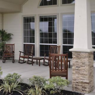 A cozy porch with wooden rocking chairs, large windows, and decorative columns surrounded by lush plants.