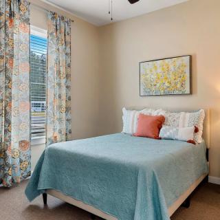 A cozy bedroom with a neatly made bed, colorful pillows, floral curtains, and a piece of artwork on the wall.