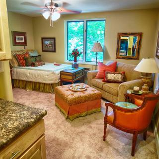 A cozy bedroom with a connected living area featuring a bed, sofa, and armchair adorned with colorful pillows. The room is warmly lit and includes large windows and tasteful decor.