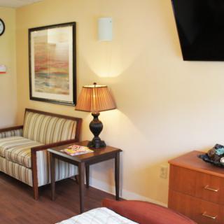 A cozy hotel room interior featuring a striped sofa, a side table with a lamp, a wall-mounted television, and artwork on the wall.