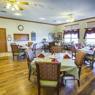 A cozy dining room with neatly set tables and chairs, featuring wooden floors and natural lighting from large windows.